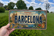 A person is holding a Barcelona license plate in front of a scenic background featuring the city's iconic landmarks and architecture.