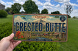 A person is holding a license plate that says "Crested Butte" in front of a scenic background with trees and mountains.