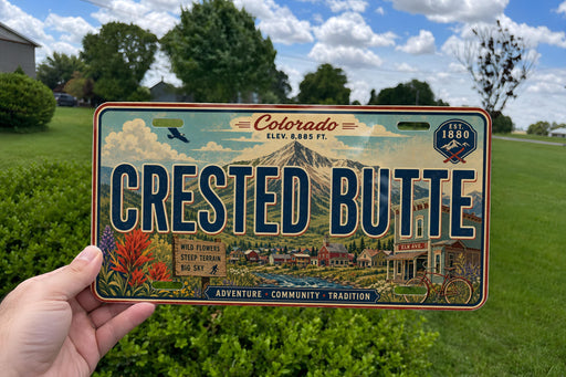 A person is holding a license plate that says "Crested Butte" in front of a scenic background with trees and mountains.