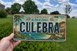 A person is holding a license plate with the word "CULEBRÁ" on it, which is a tropical island in Puerto Rico.