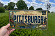 A person is holding a vintage Pittsburgh license plate with a cityscape illustration on it.