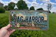 A person is holding a license plate that says "Sag Harbor" in front of a scenic background featuring a lighthouse, a sailboat, and a town.