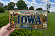 A person is holding a license plate with the word "IOWA" prominently displayed, featuring various scenes and landmarks associated with the state of Iowa.