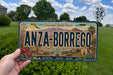 A person is holding a license plate that says "ANZA-BORREGO" in front of a desert landscape with mountains and trees.