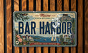 A vintage Maine license plate with the text "BAR HARBOR" and a scenic illustration of the coast, mountains, and boats.