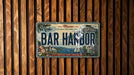 A Maine license plate with the text "BAR HARBOR" is displayed against a wooden background.
