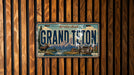 A vintage license plate from Grand Teton, with a scenic mountain landscape and moose illustration, is displayed on a wooden wall.