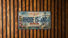 A vintage license plate from Rhode Island with a scenic illustration of the state's landmarks and a lighthouse.