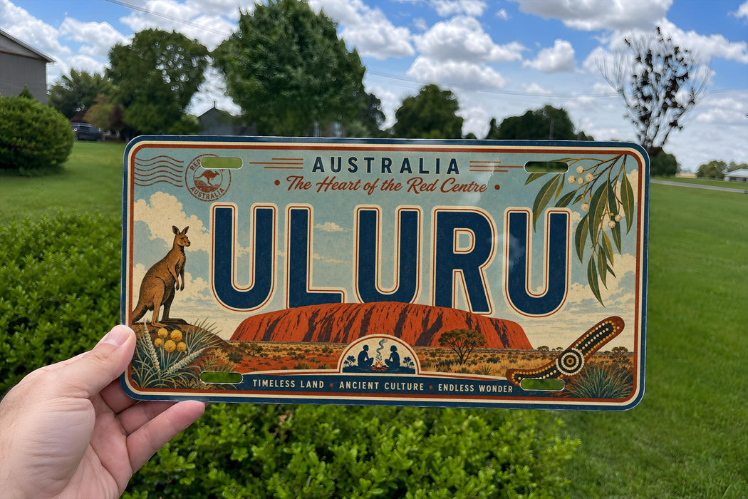 A person is holding a license plate that features the word "ULURU" prominently in the center, surrounded by various Australian landmarks and elements such as kangaroos, eucalyptus trees, and a red rock formation.