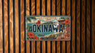 A vintage license plate from Japan with the text "OKINAWA" prominently displayed, along with various scenic elements such as a temple, ocean, and flowers.