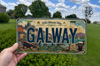 A person is holding a license plate with the word "GALWAY" prominently displayed on it. The license plate also features illustrations of a cityscape, a boat, and a glass of beer, along with the text "Wild Atlantic Way" and "Culture - Coast - Craig".