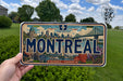 A person is holding a license plate with the word "MONTREAL" prominently displayed on it. The license plate also features a bridge and other cityscape elements, indicating it is from Quebec, Canada.
