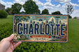 A person is holding a license plate with the text "CHARLOTTE NORTH CAROLINA City of Oaks EST. 1768" on it, with a cityscape and other elements visible on the plate.