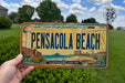 A person is holding a license plate from Pensacola Beach, Florida, which features a beach scene and the text "Pensacola Beach" prominently displayed.