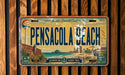 A vintage license plate from Pensacola Beach, Florida, featuring a beach scene with a lighthouse, palm trees, and a bird.