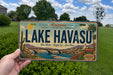A person is holding a license plate that says "Lake Havasu" with a scenic illustration of the lake and surrounding landscape.