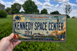 A person is holding a Florida license plate that features an illustration of the Kennedy Space Center.