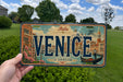 A person is holding a license plate that says "VENICE" with a cityscape of Venice on it.