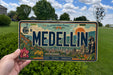 A person is holding a license plate with the word "MEDELLIN" prominently displayed on it. The license plate also features various cityscape elements, such as buildings, a bridge, and a helicopter.