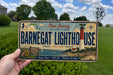 A person is holding a license plate that says "Barnegat Lighthouse Use" with a scenic background of a lighthouse and a grassy field.