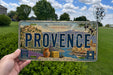 A person is holding a vintage license plate from Provence, France, which features a scenic illustration of the region.