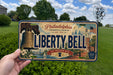 A person is holding a license plate with the image of the Liberty Bell and Philadelphia, Pennsylvania, with a clear blue sky in the background.