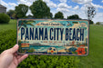 A person is holding a license plate with the text "Panama City Beach" on it, which is a tropical beach destination in Florida.