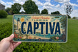 A person is holding a Florida license plate that says "CAPTIVA" in front of a scenic background featuring a lighthouse, palm trees, and a beach.