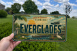 A person is holding a license plate that says "EVERGLADES" with a tropical landscape and wildlife scene on it.