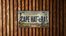 A vintage license plate from Cape Hatteras, North Carolina, with a lighthouse and coastal scenery on it, mounted on a wooden wall.