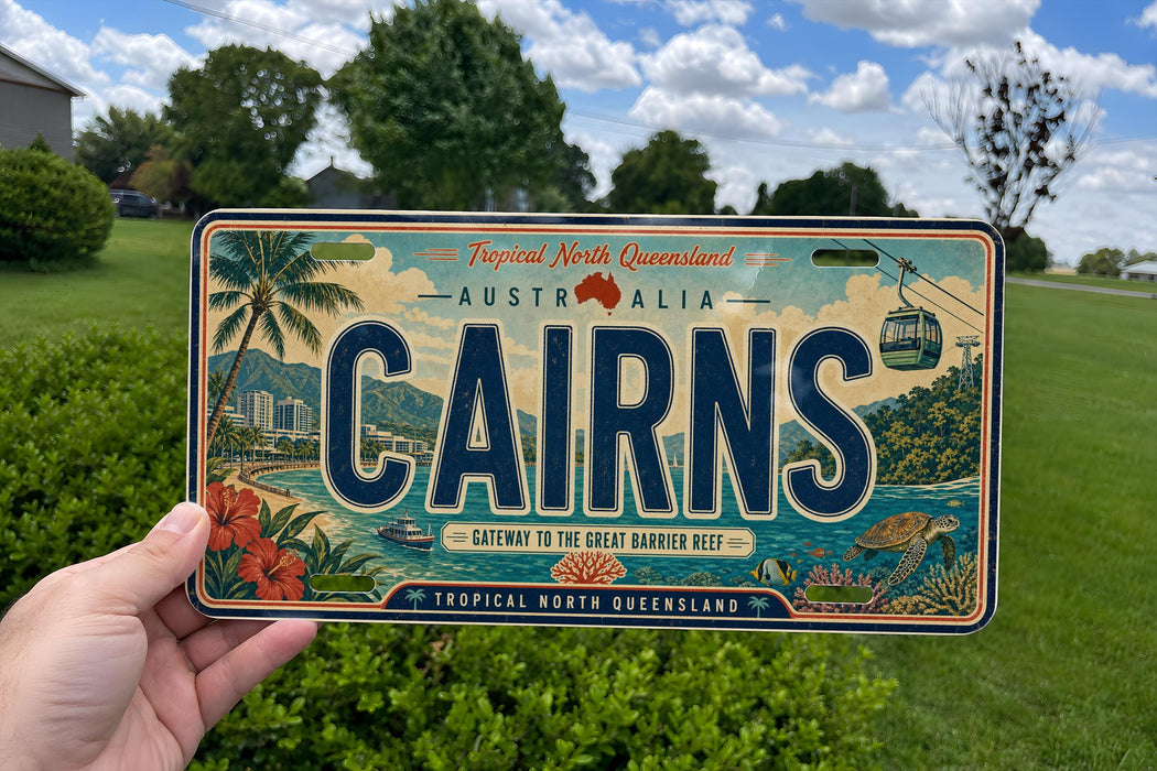 A person is holding a license plate that says "CAIRNS" in large letters, with a tropical landscape and water scene on the plate.