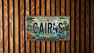 A license plate from Australia that says "CAIRNS" and is displayed against a wooden background.