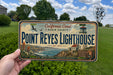 A person is holding a California Coast Marin County license plate that features the Point Reyes Lighthouse, a historic lighthouse located in Point Reyes National Seashore.