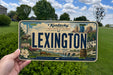 A person is holding a license plate from Kentucky that features an illustration of the city of Lexington.