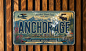 A vintage license plate from Alaska featuring a scenic mountain landscape, a moose, and a plane.