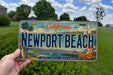 A person is holding a California license plate that says "Newport Beach" with a scenic illustration of the beach and surrounding landscape.
