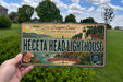 A person is holding a license plate with a scenic image of the Oregon Coast and the Heceta Head Lighthouse.