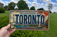 A person is holding a Toronto license plate in front of a scenic background featuring the iconic CN Tower and the Toronto skyline.