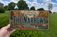 A person is holding a license plate with a scenic mountain landscape on it, with a grassy field and trees in the background.
