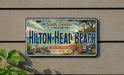 A license plate with the text "South Carolina Welcome to Hilton Head Beach" and various illustrations of landmarks and scenery, including a lighthouse, palm trees, and a beach.