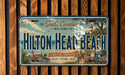 A vintage license plate with a coastal theme, featuring a lighthouse, palm trees, and a beach scene. The plate is mounted on a wooden wall.