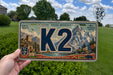 A person is holding a license plate with a mountain landscape and the text "K2" prominently displayed.