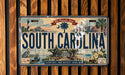 A decorative license plate from South Carolina, featuring a coastal scene with palm trees, a lighthouse, and other landmarks.