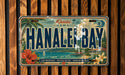 A vintage license plate from Hawaii, featuring the text "HANALEIBAN" and a tropical landscape with palm trees, flowers, and a surfer.