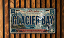A vintage license plate from Alaska featuring the name "Glacier Bay" and a scenic image of the bay with mountains, boats, and wildlife.