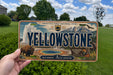 A person is holding a license plate that features the name "Yellowstone" and the park's name, along with a scenic illustration of the park.