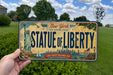 A person is holding a New York license plate that says "STATUE OF LIBERTY" with a cityscape and the Statue of Liberty in the background.