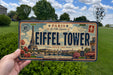 A person is holding a license plate with the Eiffel Tower and Paris in the background.