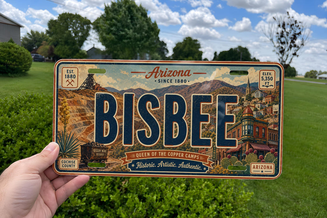 A person is holding a license plate with the word "BISBEE" prominently displayed on it. The license plate is set against a backdrop of a grassy field and trees, with a clear blue sky above.