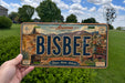A person is holding a license plate with the word "BISBEE" prominently displayed on it. The license plate is set against a backdrop of a grassy field and trees, with a clear blue sky above.